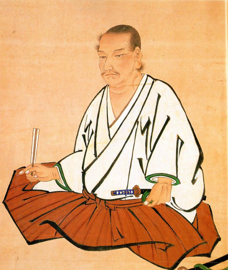 Seated portrait of Miyamoto Musashi in a white robe and brown hakama, holding a fan, at the Kumamoto Prefectural Museum of Art